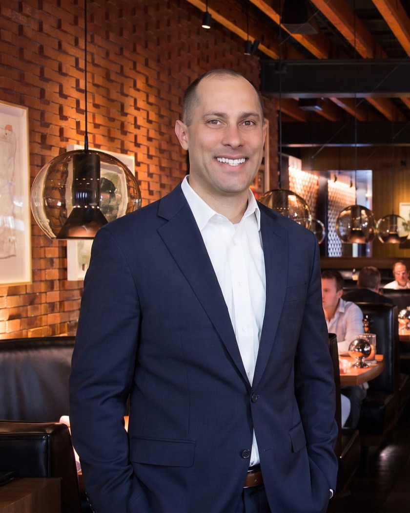 Jon Peck in a navy suit, standing in a brick-walled restaurant dining room during service