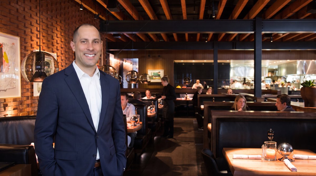 Jon Peck in a navy suit, standing in a brick-walled restaurant dining room during service, with booths and the open kitchen behind him